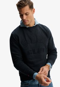 Marineblå sweatshirt med prægede logo, rund hals og ribbet kant, lagdelt over en lyseblå denimskjorte. Casual og tekstureret design.