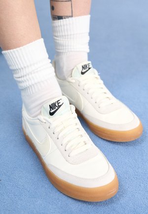 Persoon draagt witte Nike sneakers met gumzolen en witte kousen tot de kuit, staand op een lichtblauwe ondergrond, met een geometrische tatoeage op het been zichtbaar.