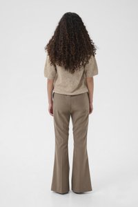 Frau mit langen lockigen Haaren steht mit dem Rücken zur Kamera, trägt einen beigen kurzärmeligen Pullover und braune weit geschnittene Hosen vor einem schlichten Hintergrund.