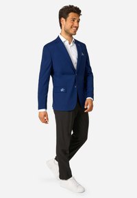 Smaidošu vīrieti, kas staigā, ģērbtu zila žakete ar izšūtu uzšuvumu, baltu krekliņu, melnas bikses un baltas čības uz neitrāla fona.