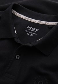 Černé polo tričko s límečkem a knoflíkem. Štítek ukazuje značku "Ombre," vyrobeno z 100% bavlny, velikost M (EUR 48).
