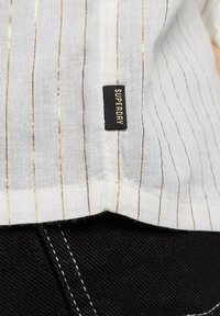 Biała tkanina z złotymi pionowymi paskami i czarną metką z napisem "SUPERDRY", przymocowaną u dołu, kontrastująca z ciemnym materiałem poniżej.