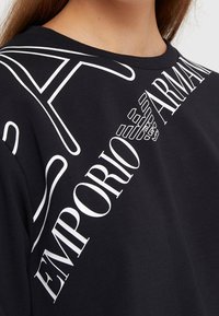 Camiseta de algod�ón negra con el prominente texto blanco "Emporio Armani" y su logo, cuello redondo y mangas cortas. Textura suave, diseño casual.