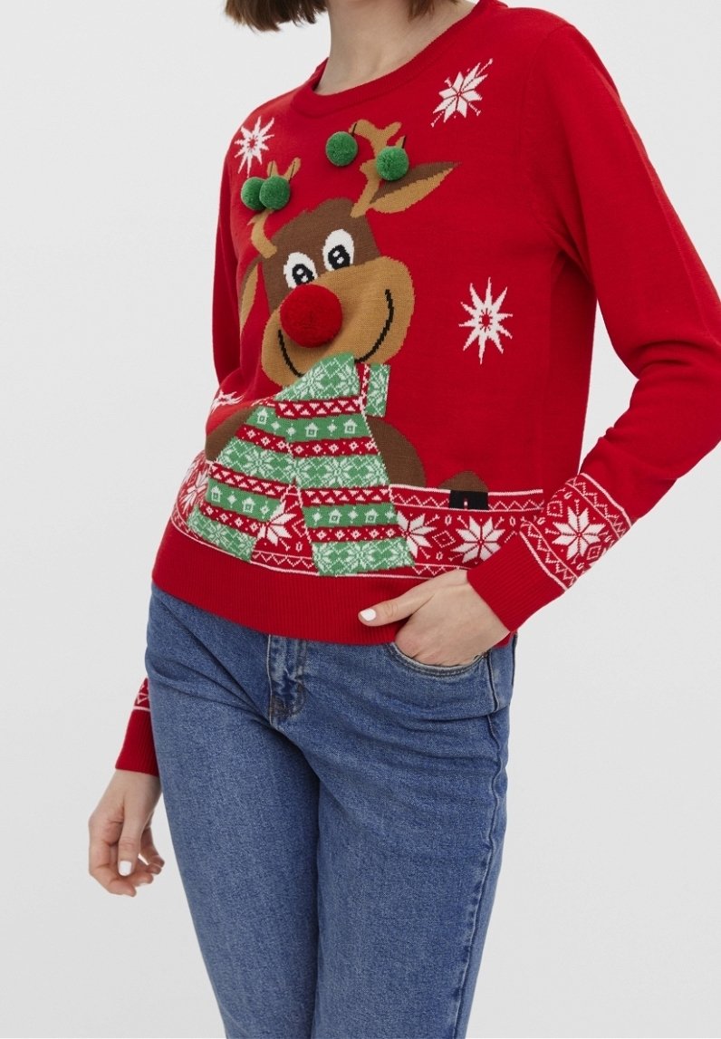 Femme portant un pull de Noël rouge avec un renne souriant, des pompons verts 3D sur les bois, un nez en pompon rouge et des motifs de flocons de neige blancs.