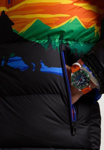 Bunte Daunenjacke mit Naturgrafiken, die grüne und orangefarbene Designs aufweist; beinhaltet einen blauen Reißverschluss. Orangefarbene Uhr mit grünem Gehäuse.