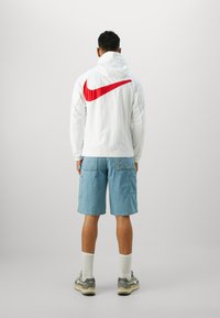 Valkoinen hupputakki, jossa on suuri punainen Nike-logo selässä, yhdistettynä sinisiin farkkushortseihin, valkoisiin sukkiin ja harmaisiin lenkkareihin.
