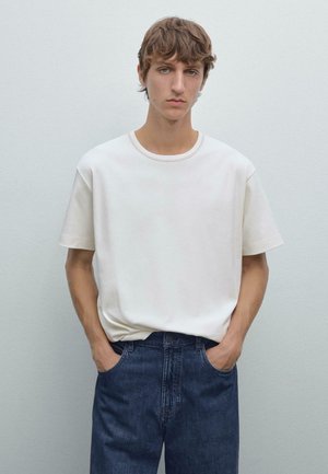 Weißes Baumwoll-T-Shirt mit kurzen Ärmeln und rundem Ausschnitt, kombiniert mit blauen Jeans. Das Model steht mit den Händen in den Taschen.