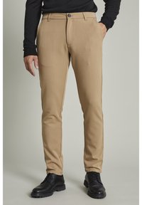 Beżowe spodnie o wąskim kroju, wykonane z gładkiego materiału, z zapięciem na guzik i kieszeniami. Nosi się je z czarnymi butami i czarnym swetrem.