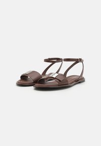 Brune lærsandaler med åben tå, der har en bred rem med en sølv hardware detalje og en slank ankelrem. Flad sål.