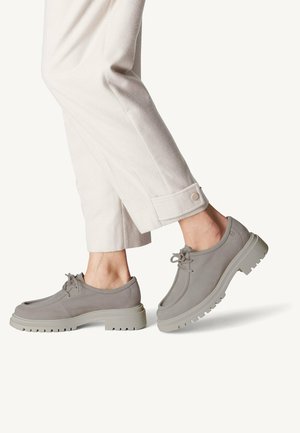 Personne portant un pantalon beige clair et des chaussures à lacets grises à semelle épaisse, un pied levé dans un mouvement de marche sur un fond blanc.