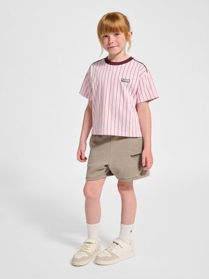 Junges Mädchen mit roten Haaren trägt ein rosa gestreiftes T-Shirt, beige Shorts, weiße Socken und beige Turnschuhe, steht vor einem einfarbigen Hintergrund.