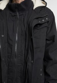 Chaqueta negra con cuello alto, cierre frontal con cremallera y botones a presión. Cuenta con un forro suave y texturizado y múltiples bolsillos para almacenamiento.
