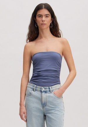 Frau mit langen dunklen Haaren trägt ein trägerloses blaues gerafftes Top und hellblaue Jeans, steht mit einer Hand in der Tasche vor schlichtem Hintergrund.