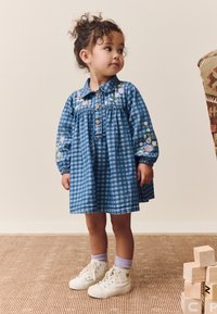 Mėlyna ginghamo suknelė su mygtukų užsegimu, siuvinėtais gėlėmis ant pečių ir pūstomis rankovėmis. Derinama su baltais aukštu aulo sportbačiais ir violetinėmis kojinėmis.