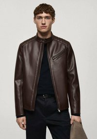 Veste en cuir marron avec un col montant, poche poitrine zippée, design épuré et détails de couture ; portée sur une chemise noire.