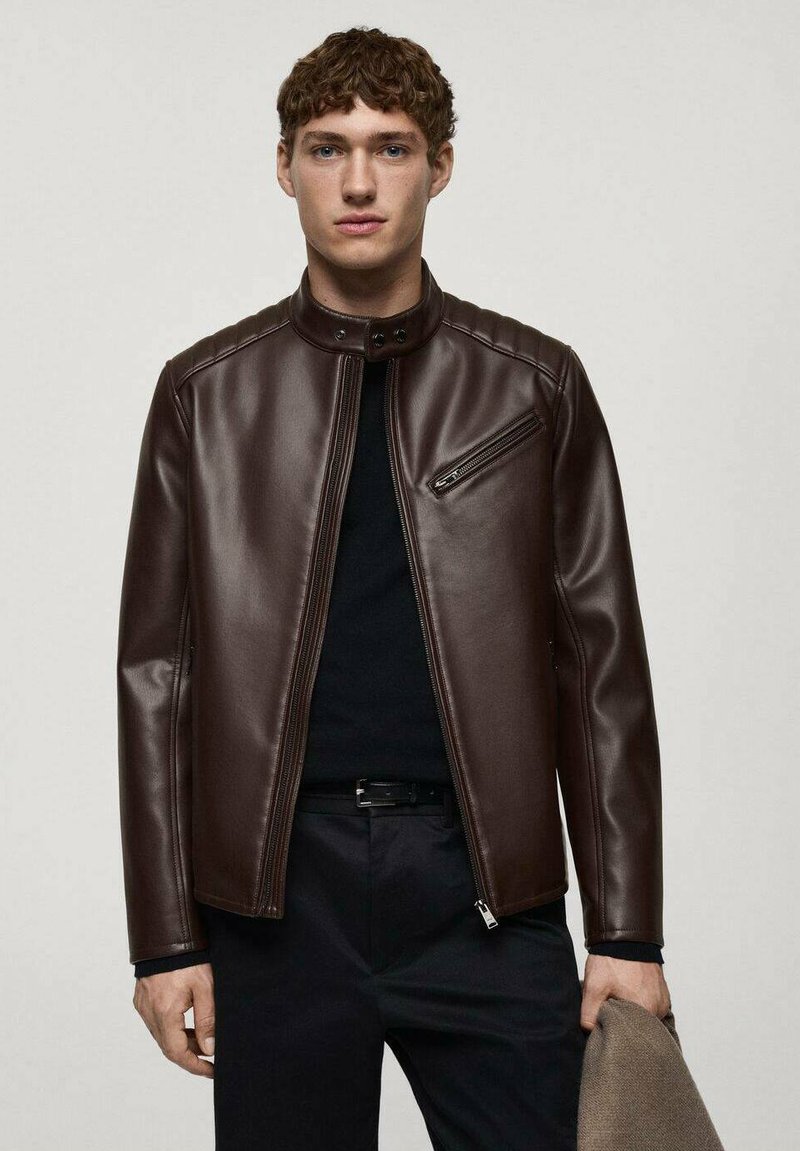 Veste en cuir marron avec un col montant, poche poitrine zippée, design épuré et détails de couture ; portée sur une chemise noire.