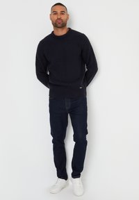 Dunkelblauer Strickpullover mit strukturiertem Muster, Rundhalsausschnitt. Kombiniert mit dunklen Jeans und weißen Sneaker. Das Model steht mit gefalteten Händen.