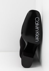 Semelle de chaussure à talon haut noire vue de dessous avec le nom de la marque « Calvin Klein » imprimé en blanc, sur fond blanc.