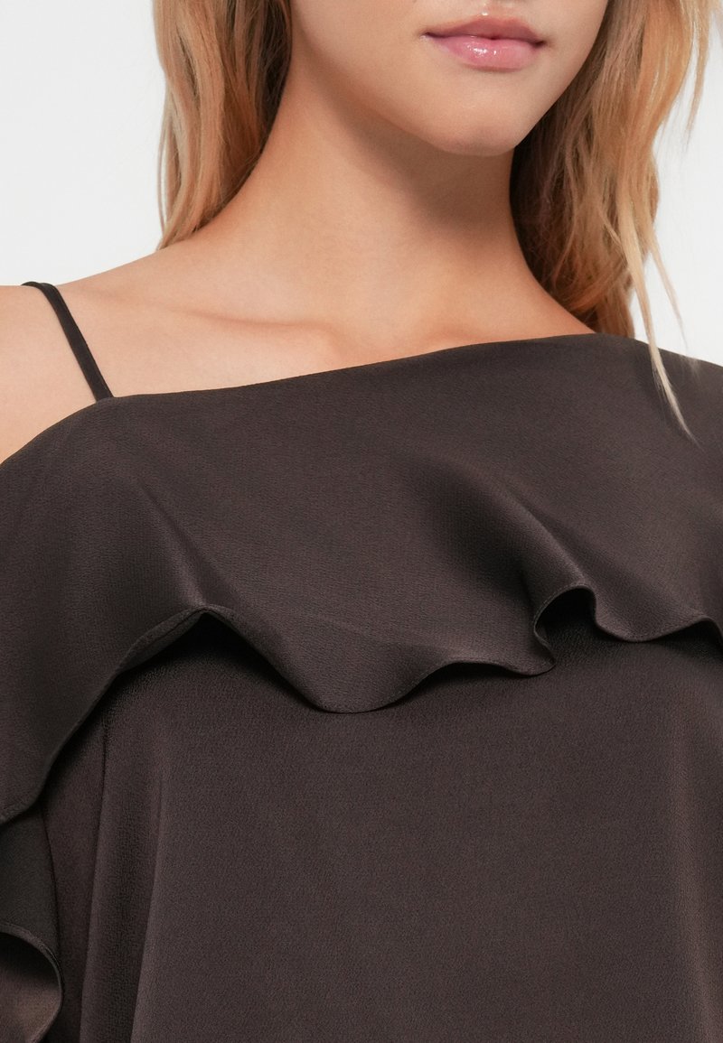 Femme portant un haut noir décolleté avec un col froncé et une fine bretelle, mettant en valeur ses lèvres et son collarbone.