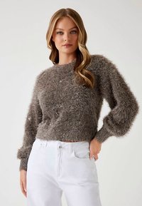 Rudas pūkuotas megztinis su apvalia iškirpte, ilgomis pūstomis rankovėmis ir trumpu kraštu. Derinamas su baltais džinsais. Audinys atrodo minkštas ir tekstūruotas.