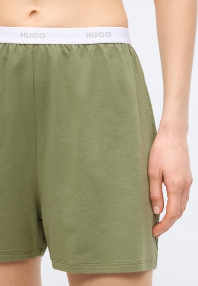 Vue de la taille jusqu'à mi-cuisse d'une personne portant un short vert olive avec une ceinture élastique blanche portant la marque "HUGO" sur un fond clair.