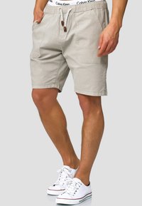 Shorts en coton beige avec une taille à cordon, poches avant et poche arrière, associés à des baskets blanches avec un accent rouge.