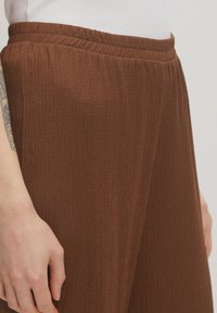 Bruine wijde broek met een ribbelstructuur en een elastische tailleband, zijaanzicht met een hand die tegen de stof leunt.