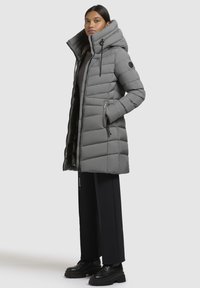 Woman standing with hands in pockets wearing long gray quilted puffer coat, black pants, and black chunky shoes against a plain background.