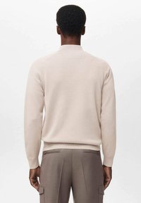 Beige knit sweater with long sleeves, ribbed cuffs and hem, round collar, and smooth texture, worn over brown trousers.