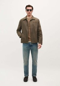 Olijfgroene, knoop-jack met borstzak over een tan kleurige gebreide trui en lichtblauwe jeans. Zwarte schoenen en een zonnebril maken de look compleet.