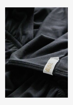 Schwarzer Stoff mit glatter Textur und leichtem Stretch; verfügt über ein weißes Etikett mit goldener Schrift, das in die Naht eingestickt ist.