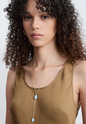 Kobieta o kręconych ciemnych włosach nosi złote kolczyki koła, złoty naszyjnik z dwoma jasnoniebieskimi kamieniami oraz brązowy top bez rękawów.