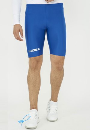 Uomo che indossa pantaloncini atletici compressivi Legea blu e sneakers bianche, in piedi su uno sfondo bianco neutro.