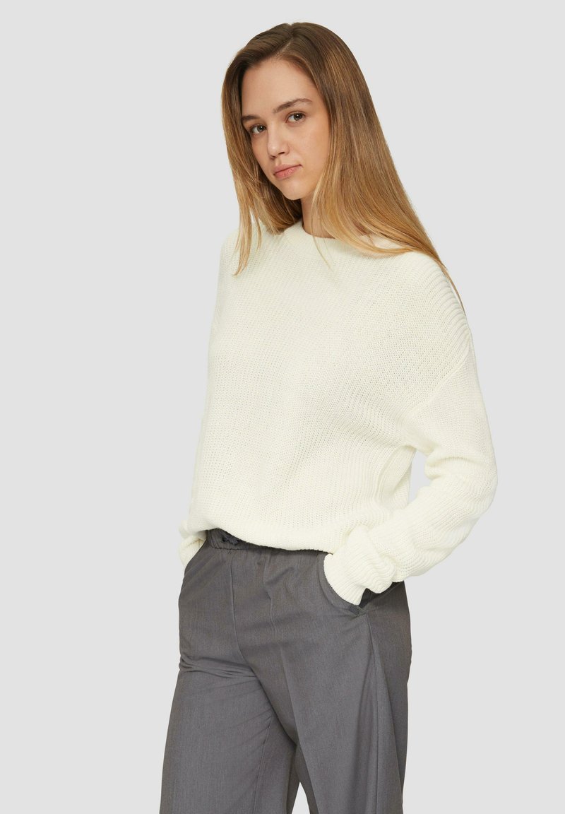 Witte gebreide trui met een ontspannen pasvorm en geribde manchetten, gecombineerd met grijze broek. De trui heeft een gestructureerde afwerking en een ronde hals.