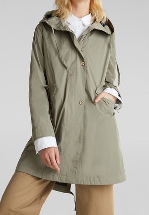 Femme portant un imperméable à capuche vert olive clair avec des boutons à l'avant, des poignets de chemise blancs visibles, et un pantalon beige, main dans la poche.