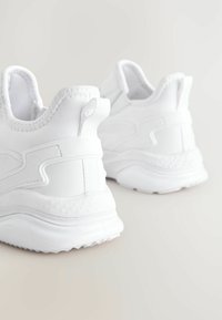 Baskets blanches de sport avec un dessus en synthétique lisse, des accents texturés, une semelle rembourrée et une languette au talon pour un enfilage facile.