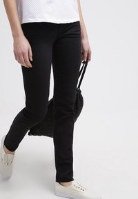 Černé slim-fit kalhoty s hladkou texturou, doplněné bílými slip-on botami s kovovými akcenty. V jedné ruce se drží prošívaná černá taška.