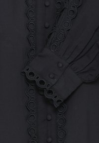 Gros plan d'un chemisier noir avec des boutons ronds en tissu, une bordure en dentelle festonnée et une manche bouffante avec un poignet brodé.