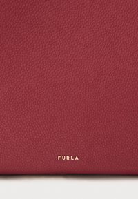 Sarkana teksturēta ādas soma ar gludu virsmu. Apakšējā labajā stūrī redzams izteiksmīgs zelta logotips "FURLA". Vienkāršs un elegants dizains.