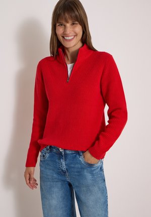 Femme souriante portant un pull rouge zippé et un jean bleu, debout avec une main dans la poche devant un fond clair uni.