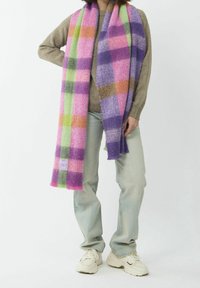 Personne portant un pull beige, un jean bleu clair, des baskets blanches et une longue écharpe à carreaux colorée avec des tons rose, violet, vert et orange.