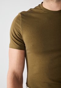Olivenfarget t-skjorte med korte ermer laget av mykt, glatt stoff. Rund hals med en tettsittende design og rene sømdetaljer på ermene.