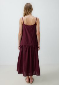 Abito lungo bordeaux con spalline sottili, gonna a strati e una superficie in cotone morbido. Presenta un corsetto aderente e una parte inferiore fluente.