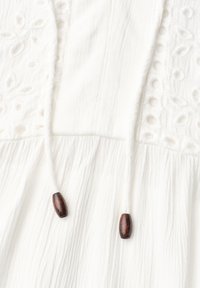 Tissu blanc texturé avec broderie florale, avec deux cordons blancs suspendus avec des perles en bois foncé aux extrémités.