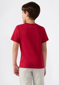 Junge, der mit dem Rücken zur Kamera steht, trägt ein schlichtes rotes Kurzarmhemd und hellgraue Hose vor einem weißen Hintergrund.