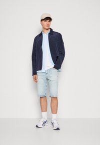Giacca leggera blu navy sopra una camicia azzurro chiaro con bottoni, abbinata a pantaloni pantaloni di jeans chiari e sneakers bianche con dettagli blu.
