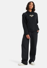 Sudadera negra con bolsillo frontal y cordón, con un logo dorado. Combinada con pantalones de chándal negros holgados y zapatos blancos.