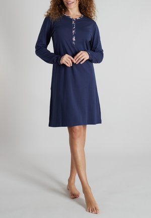 Frau trägt ein marineblaues, knielanges Kleid mit langen Ärmeln und Blumenverzierung am Ausschnitt und an der Knopfleiste, steht barfuß.