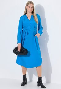 Sininen pitkähihainen mekko, jossa on napitettu kaula-aukko, vyötäröllä vyö ja sivutaskut, yhdistettynä mustaan laukkuun ja nilkkureihin. Liukas pinta.