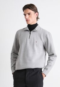 Pilka užtrauktuku užsegama megztinis iš minkštos medžiagos, su aukšta apykakle ir mažuoju logotipu ant krūtinės. Su juodomis kelnėmis.